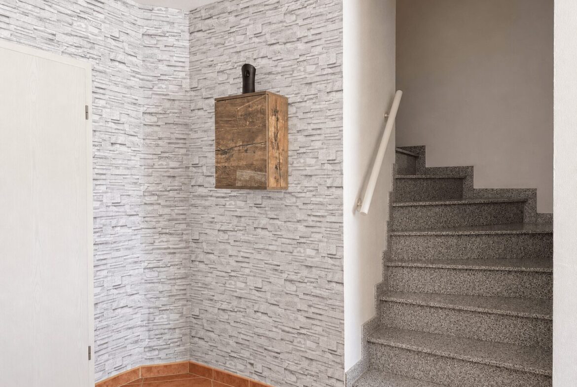 Interior stairwell with gray textured stone walls, a small wood wall cabinet, and a gray terrazzo staircase with a white handrail