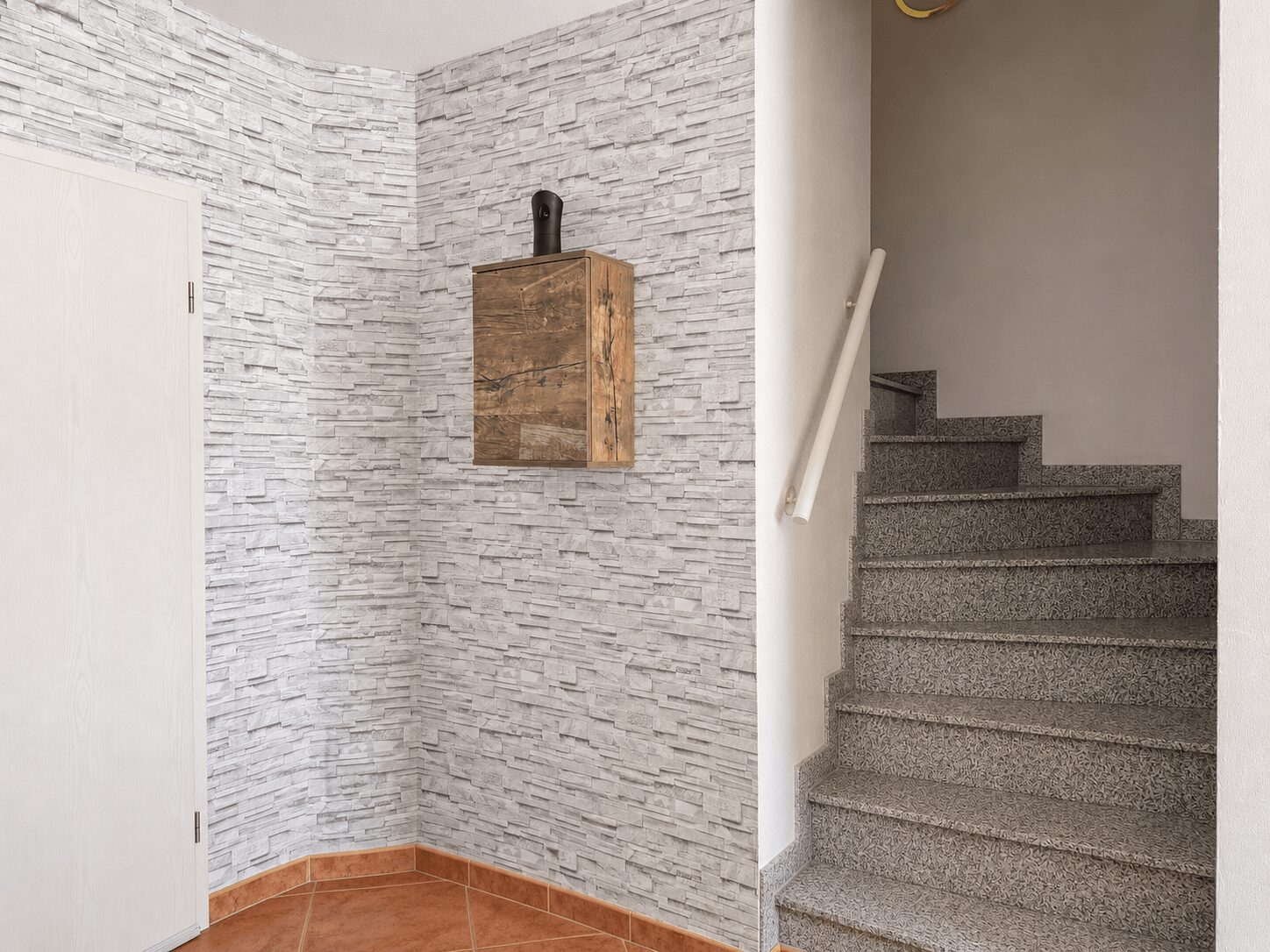 Interior stairwell with gray textured stone walls, a small wood wall cabinet, and a gray terrazzo staircase with a white handrail