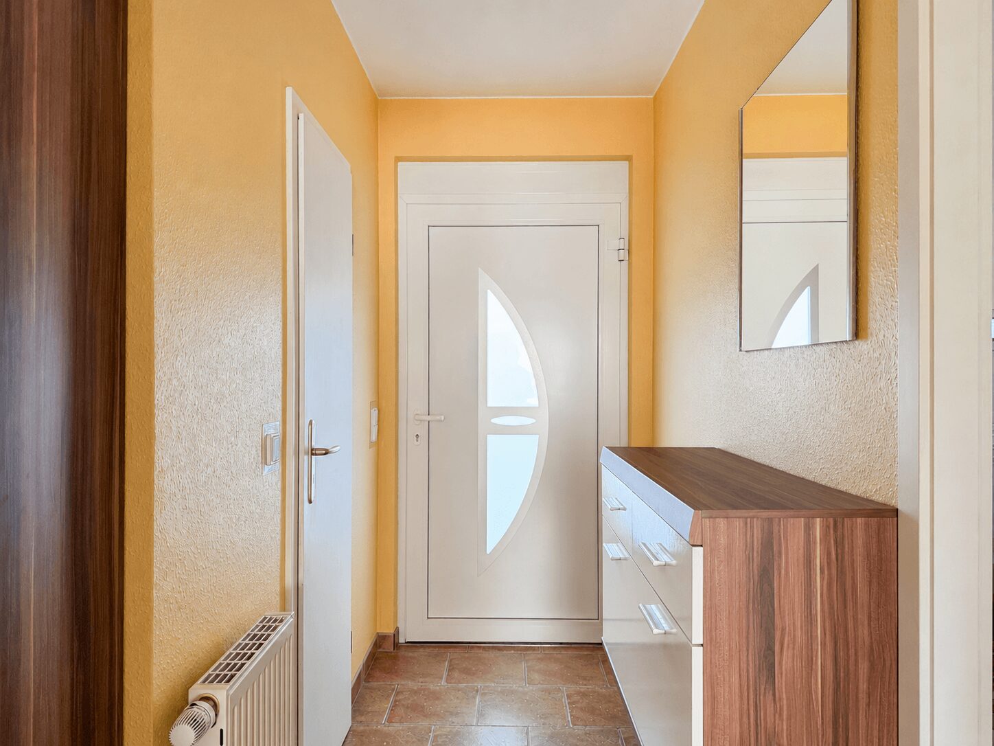 Bright entry hallway with yellow walls, leading to a white front door that has a curved window insert; a wooden-and-white console cabinet is on the right with a wall mirror above.