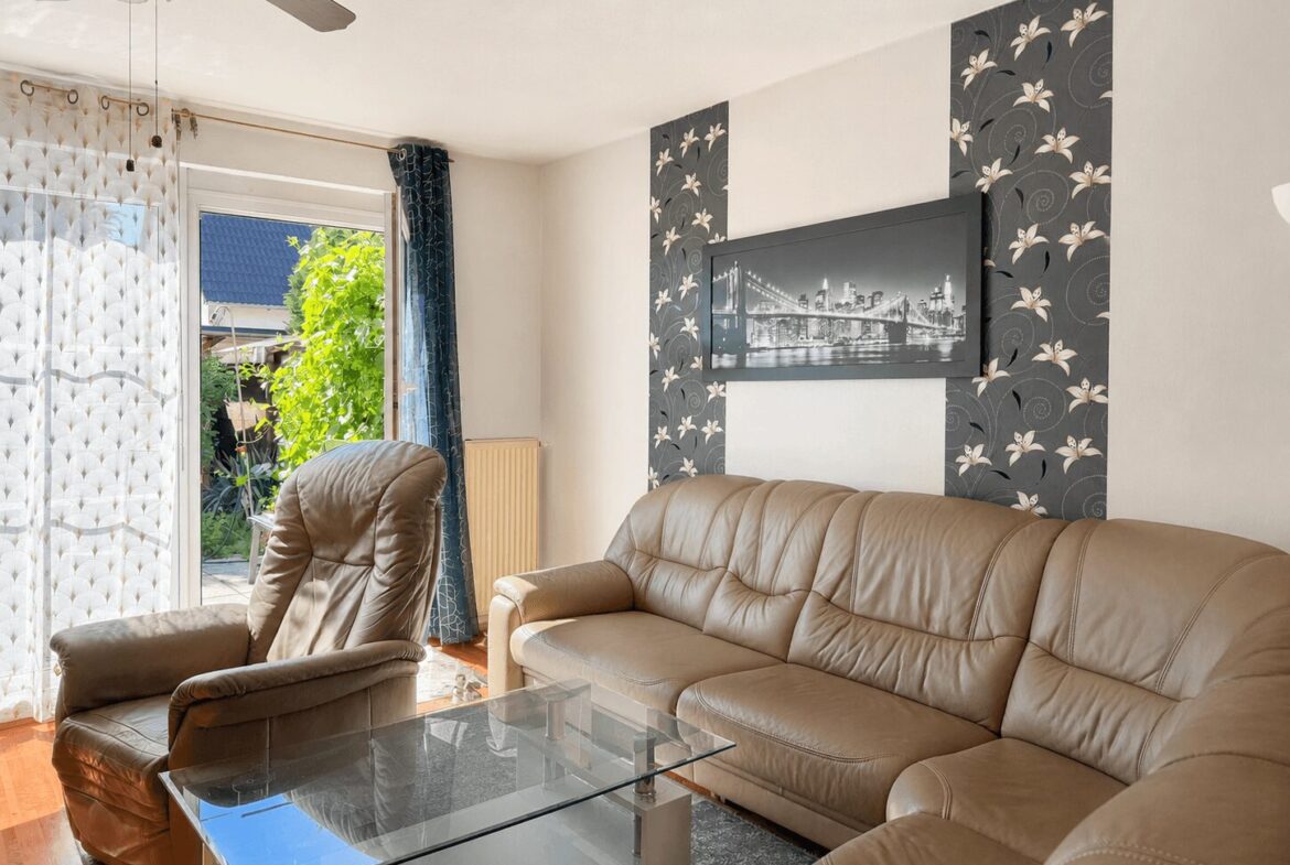 Living room with beige leather sofa, matching armchair, and a glass coffee table; large window to a sunny outdoor area with greenery and sheer patterned curtains behind the seating area.