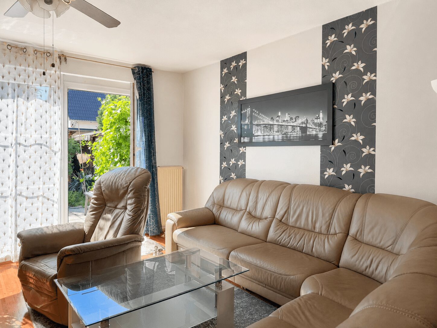 Living room with beige leather sofa, matching armchair, and a glass coffee table; large window to a sunny outdoor area with greenery and sheer patterned curtains behind the seating area.