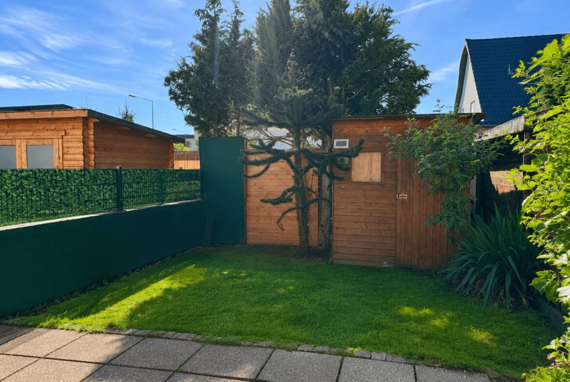 Backyard with a small wooden shed and a taller wooden structure, green lawn, and paved path under a bright blue sky.