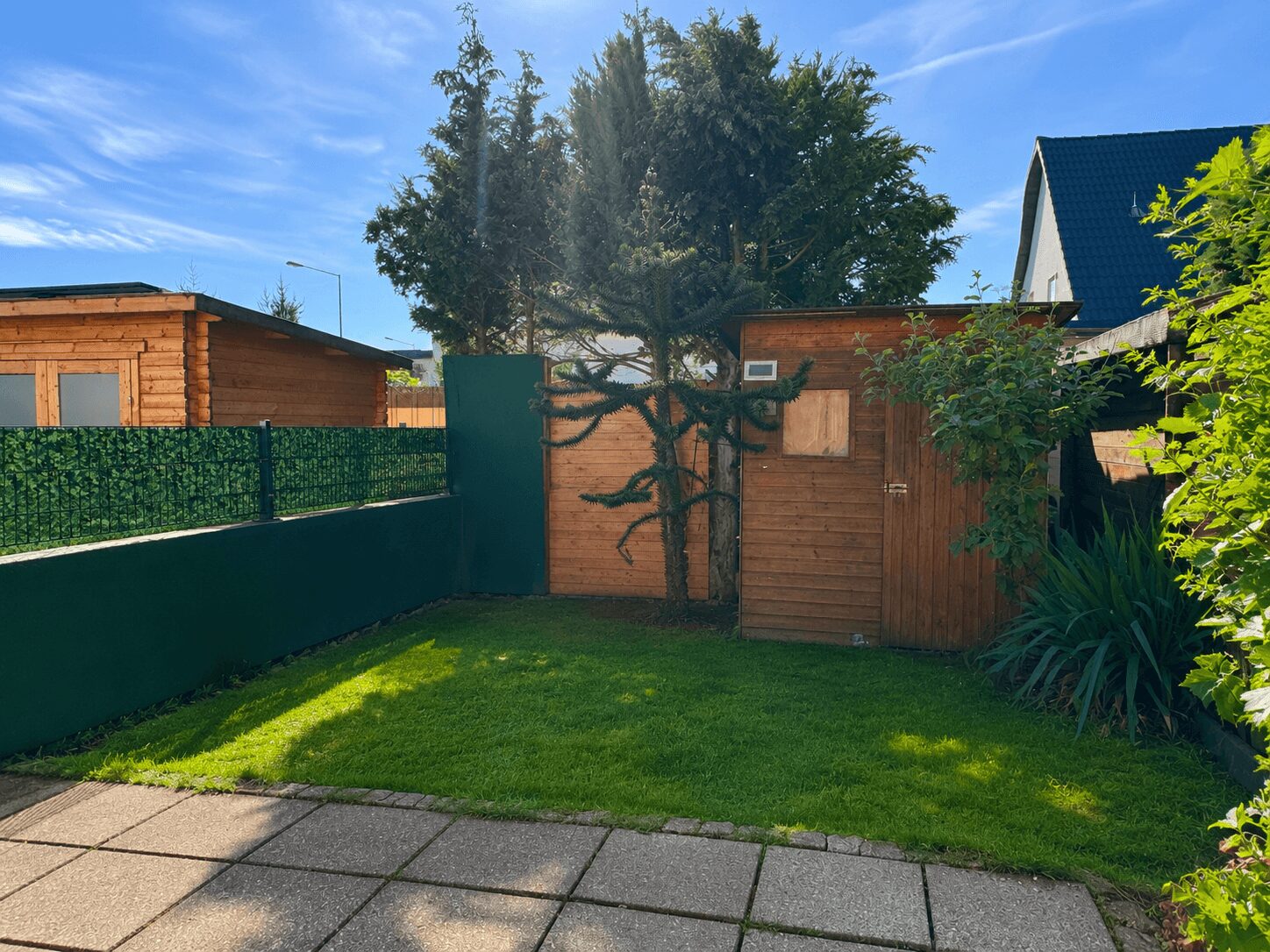 Backyard with a small wooden shed and a taller wooden structure, green lawn, and paved path under a bright blue sky.