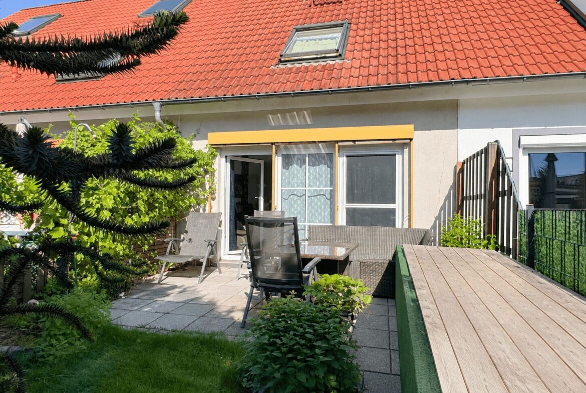 Backyard patio of a house with a red tile roof, yellow awning, glass doors, and wicker seating amid greenery on a tiled deck.