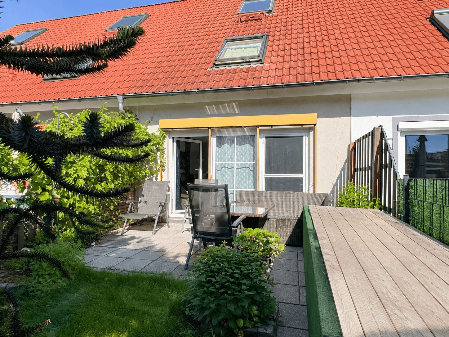 Backyard patio of a house with a red tile roof, yellow awning, glass doors, and wicker seating amid greenery on a tiled deck.