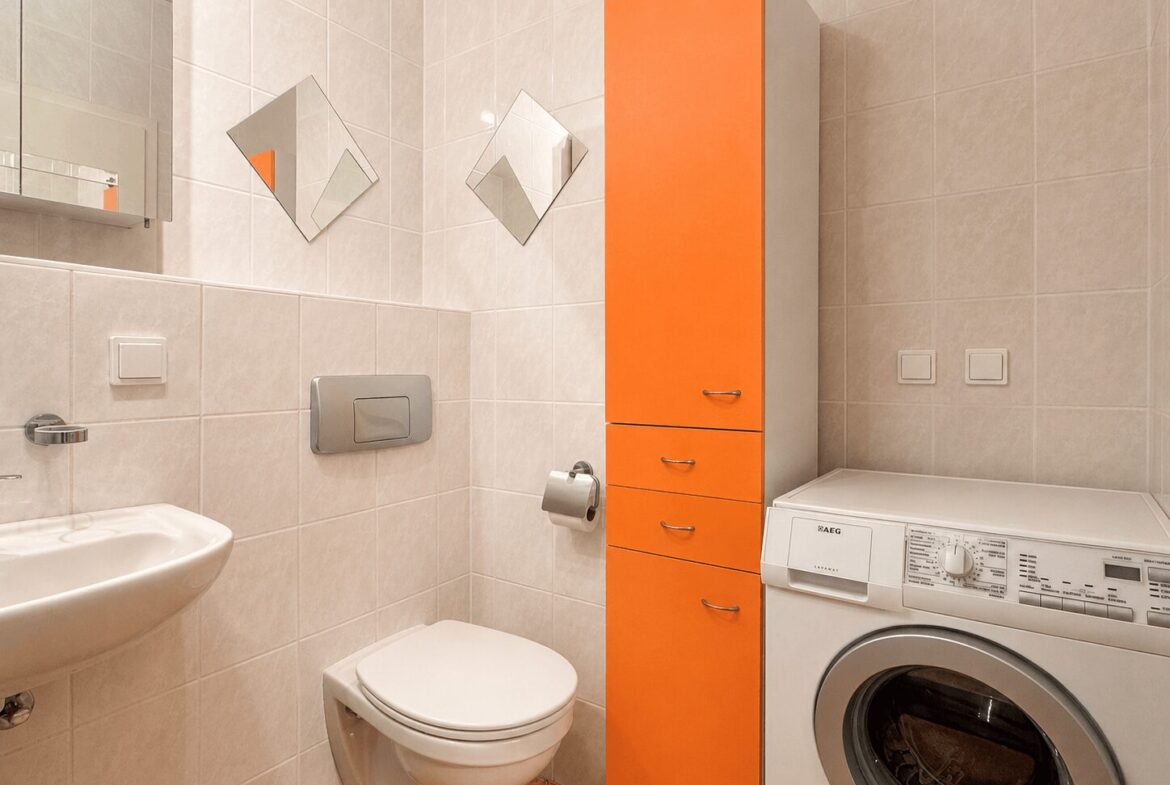 Modern bathroom with white tiles, an orange tall storage cabinet, toilet, sink, and a front-loading washing machine.