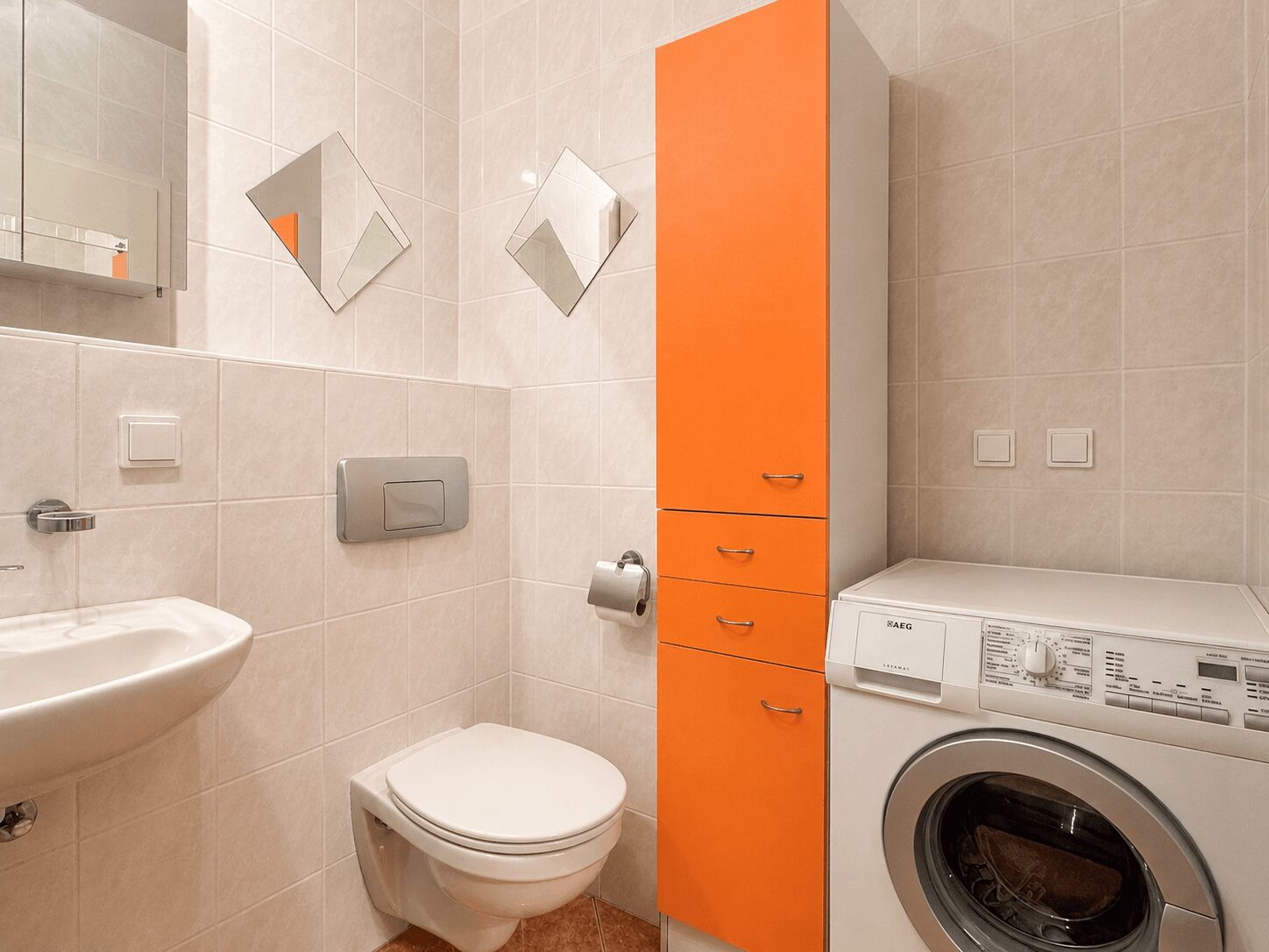 Modern bathroom with white tiles, an orange tall storage cabinet, toilet, sink, and a front-loading washing machine.