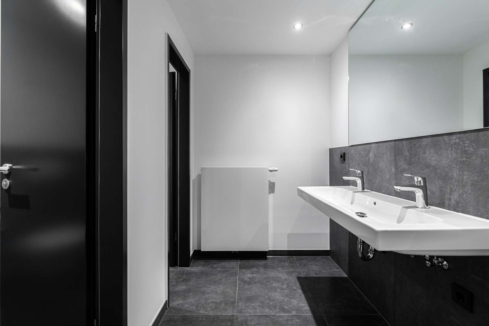 Modern bathroom with dual rectangular sinks and chrome faucets, dark tiled wall, and a large mirror on the right edge of the image.