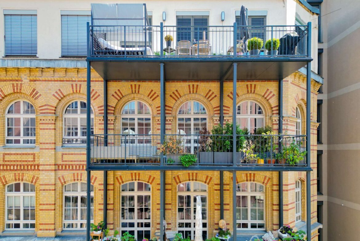 Brick apartment facade with arched windows and a blue metal balcony filled with potted plants and outdoor furniture.
