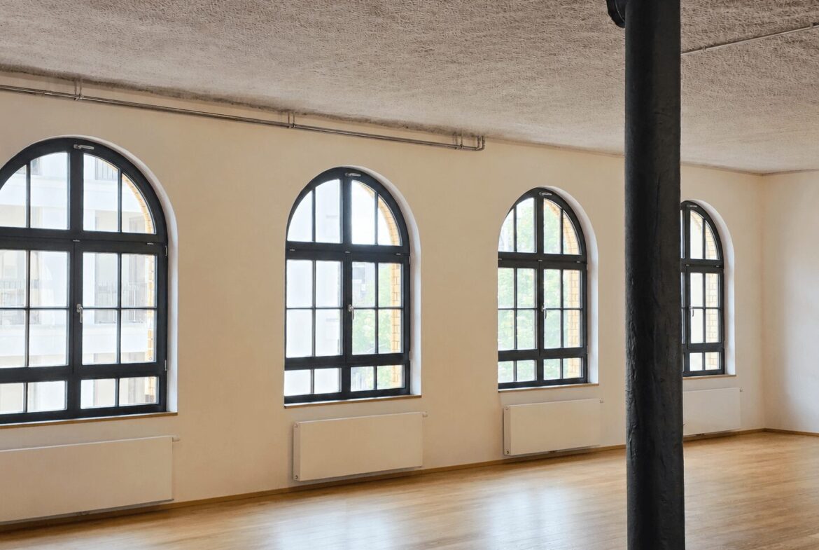 Empty loft-style room with four large arched windows along the wall, a dark column in the foreground, and a light wood floor with radiators beneath the windows.