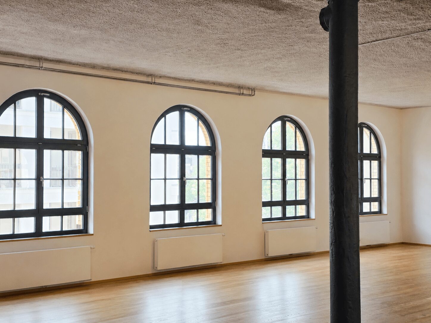 Empty loft-style room with four large arched windows along the wall, a dark column in the foreground, and a light wood floor with radiators beneath the windows.