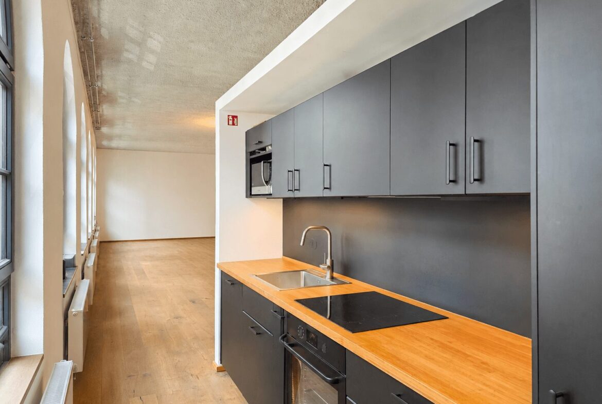 Long kitchen with gray cabinets on the right, wooden counter, sink and stovetop, and large windows along the left wall.