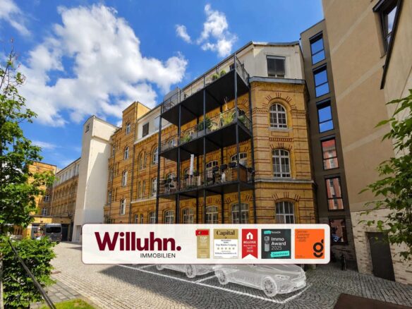 Brick building with black metal balconies advertising Willuhn Immobilien on a white banner in the foreground, blue sky above.