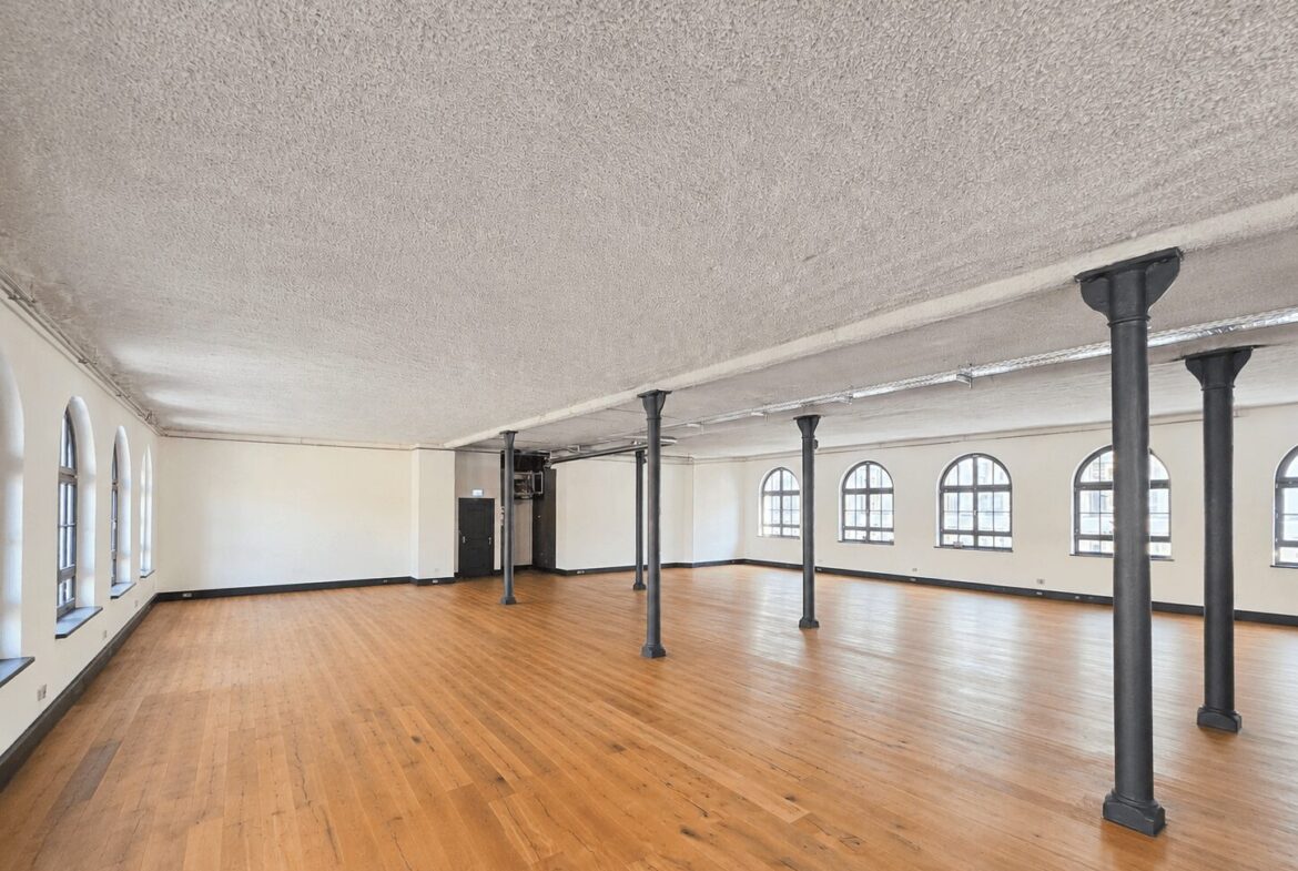 Large empty studio or event hall with wooden floor, white walls, and black support columns, lined with arched windows along the right wall