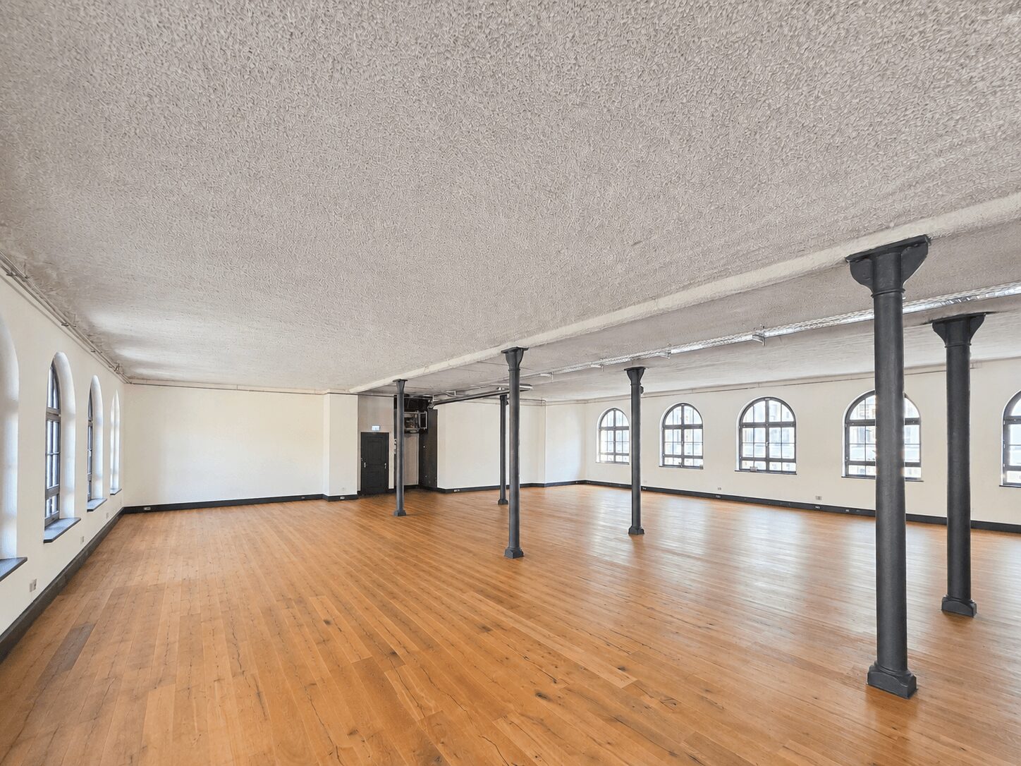 Large empty studio or event hall with wooden floor, white walls, and black support columns, lined with arched windows along the right wall