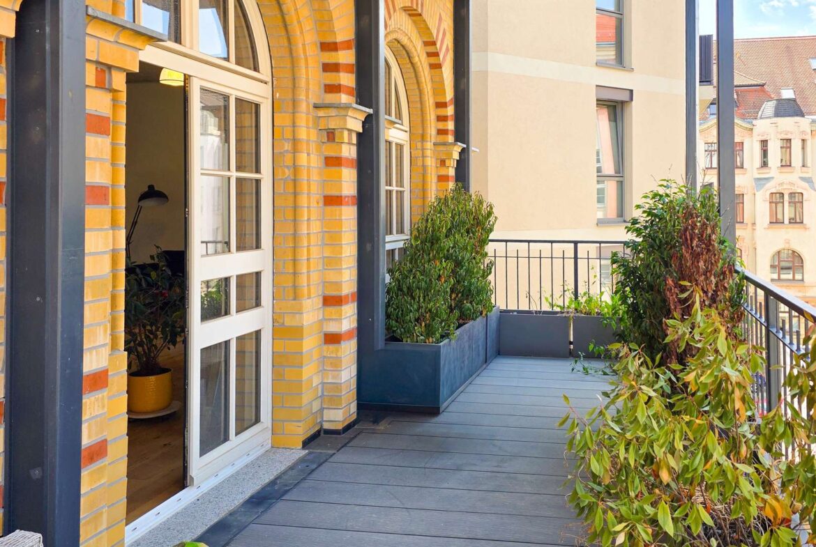 Open glass French doors onto a sunny balcony with yellow brick arches, blue-gray decking, and rectangular planters with leafy plants.