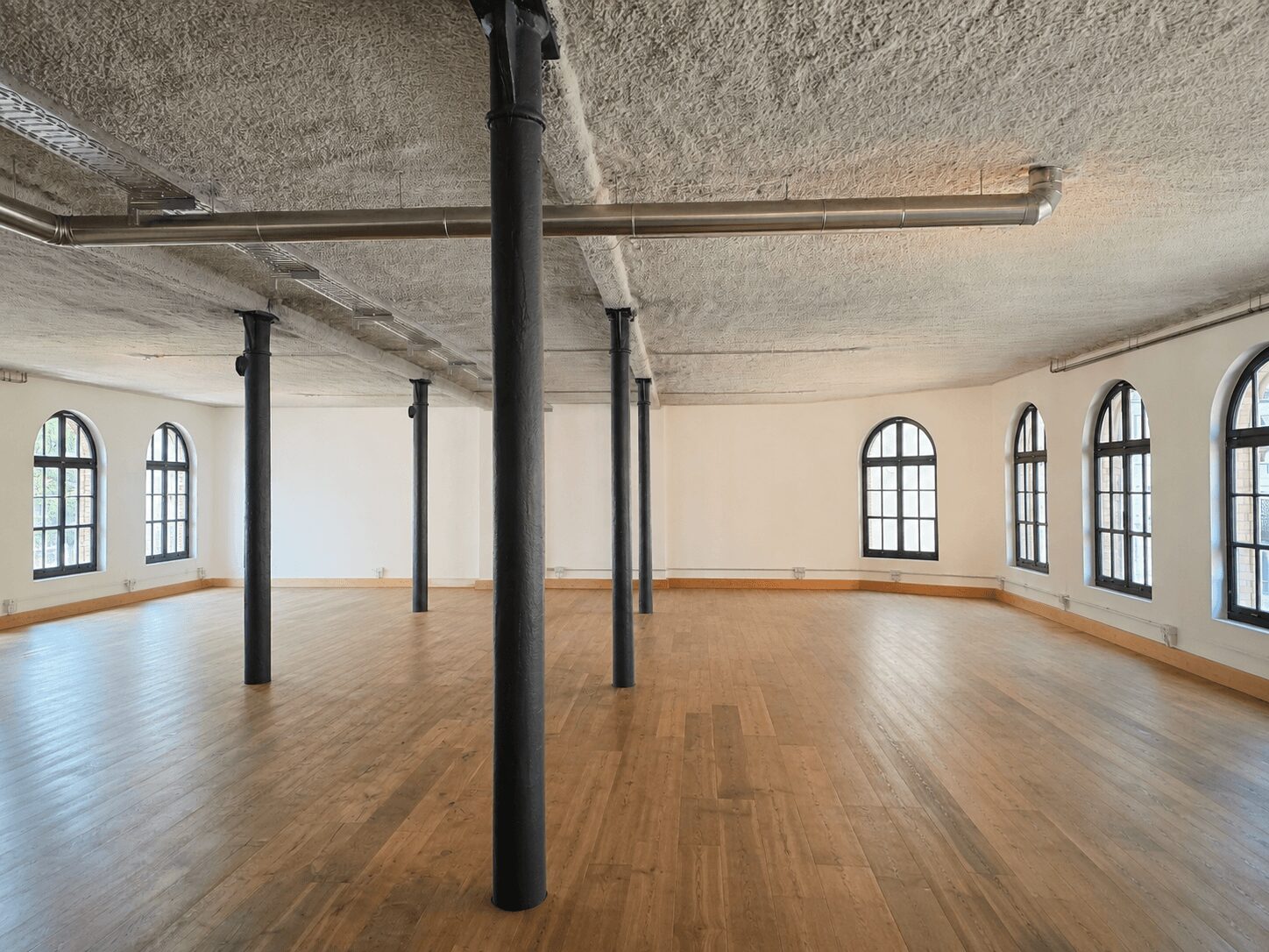 Empty studio room with wooden floor, white walls, and black circular columns supporting an industrial ceiling with exposed pipes and ducts.