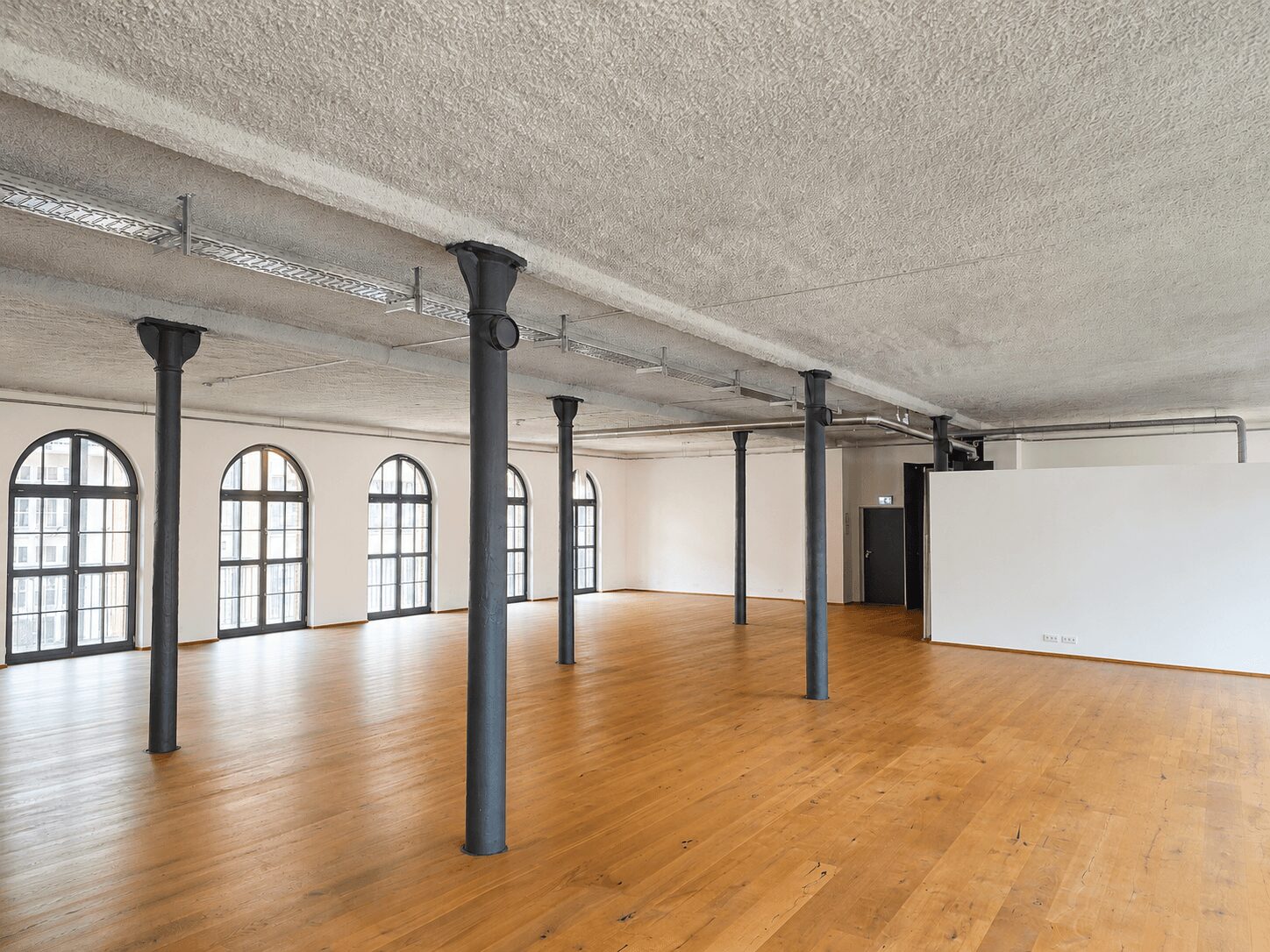 Industrial loft with tall arched windows, black columns, and a warm wooden floor.