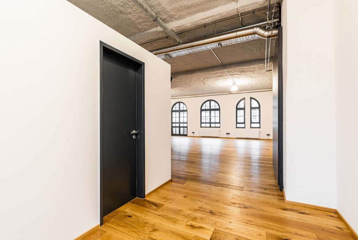 Bright hallway opening into a large room with arched windows, wooden floor, and exposed ceiling pipes.