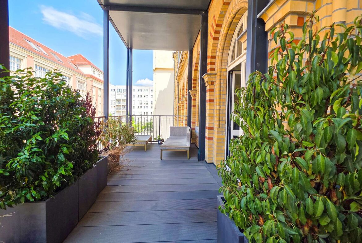 Urban balcony with a wooden deck, tall planters and green shrubs along the sides, two lounge chairs, brick building facade on the right, and a cityscape in the distance under a blue sky.
