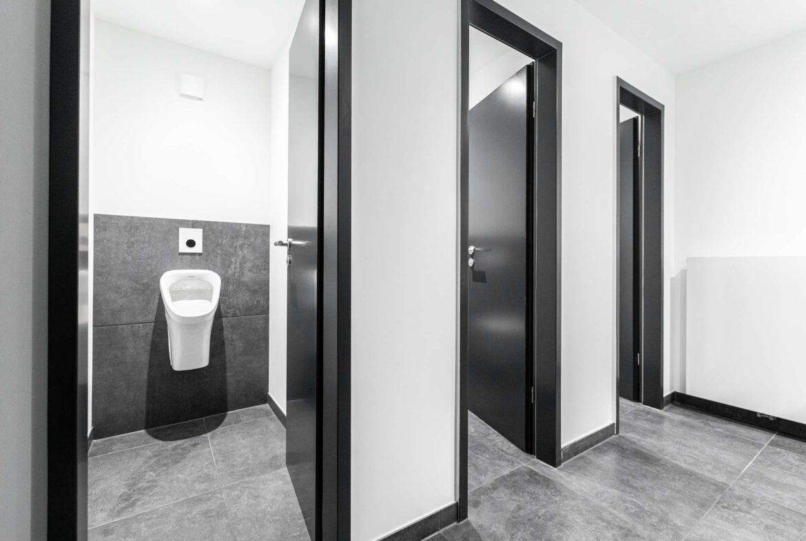 Public restroom corridor with dark-framed stall doors; a wall-mounted urinal is visible in the first stall.
