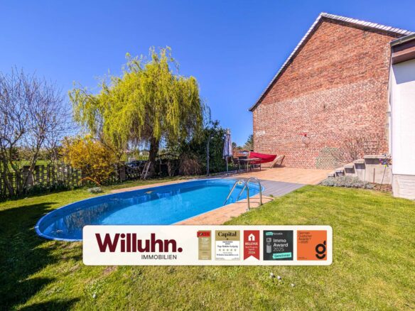 Backyard with a curved blue swimming pool, grassy lawn, and a brick house wall; a large weeping tree and leafless trees line the yard under a clear blue sky; foreground shows a white sign with Willuhn Immobilien and award stickers.