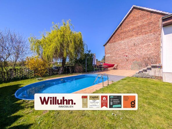 Backyard with a curved blue swimming pool, grassy lawn, and a brick house wall; a large weeping tree and leafless trees line the yard under a clear blue sky; foreground shows a white sign with Willuhn Immobilien and award stickers.