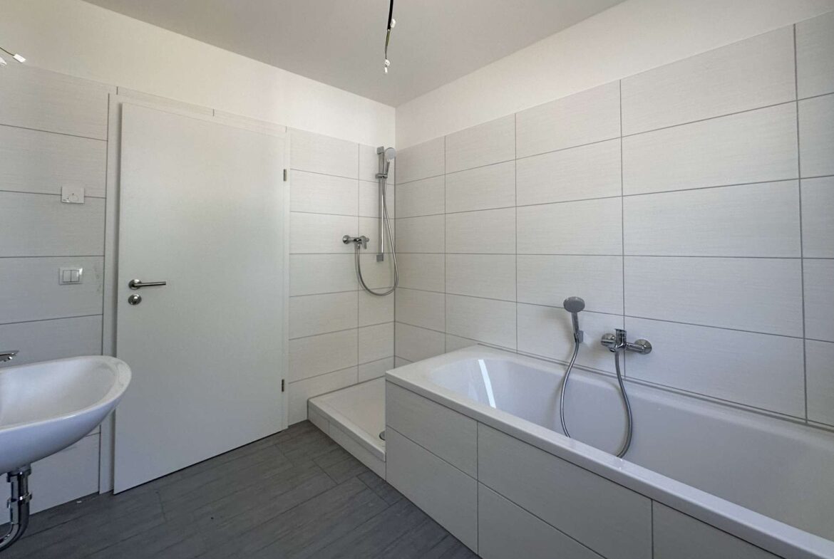 Modern white bathroom with a built-in bathtub, handheld shower, and white sink against tiled walls.