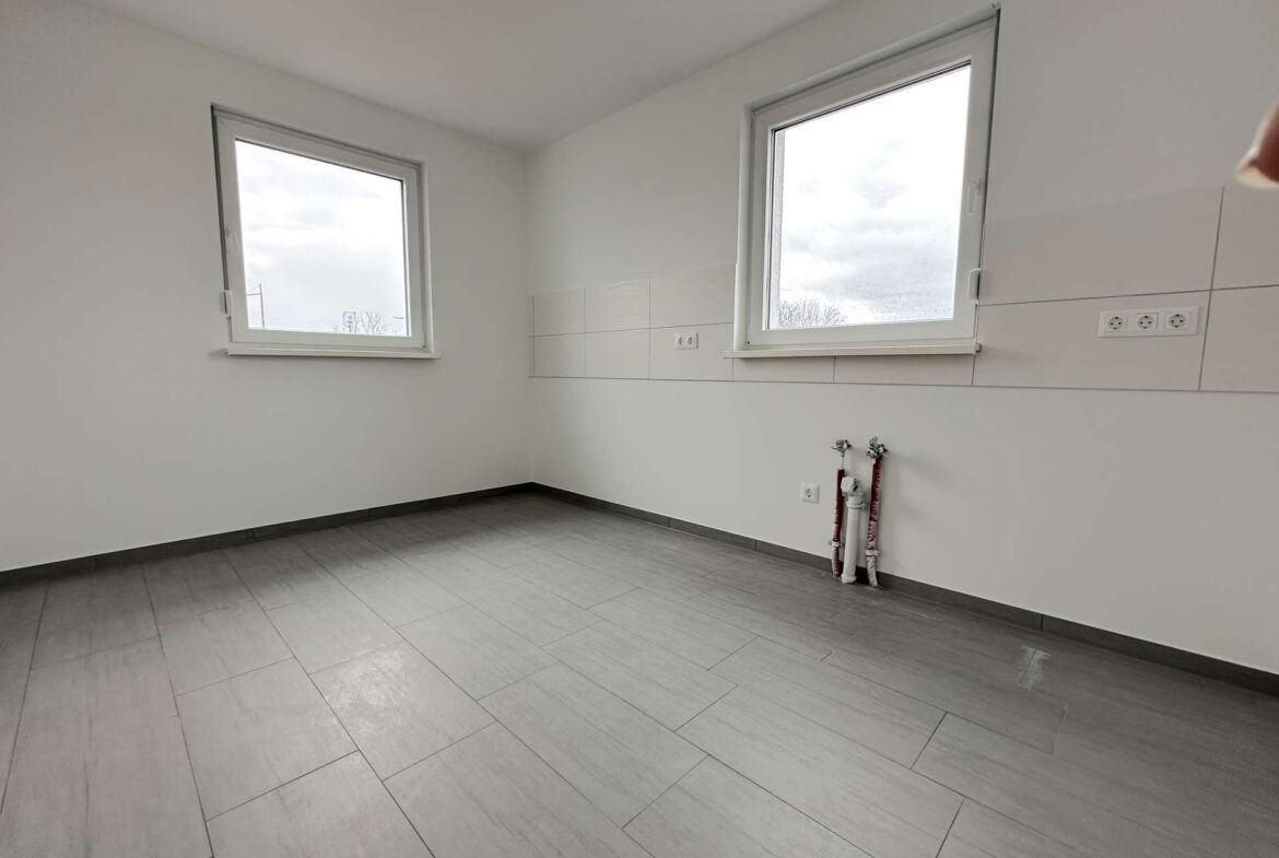 Empty white utility/room with two large windows and exposed plumbing pipes along the wall.