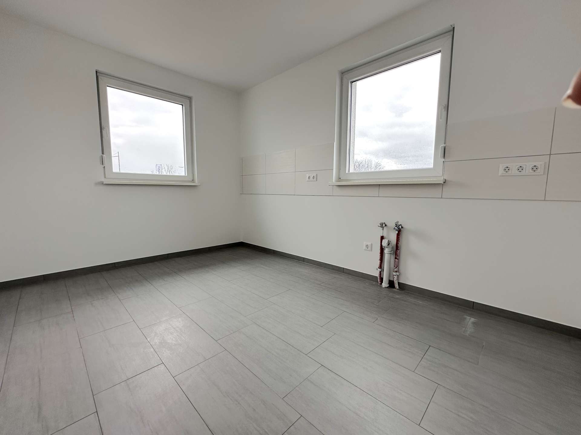 Empty white utility/room with two large windows and exposed plumbing pipes along the wall.