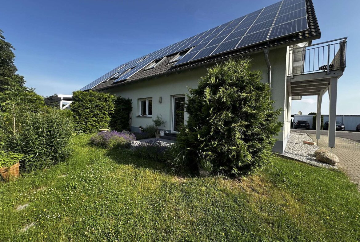 Residential house with a large array of solar panels on the sloped roof, front garden and driveway visible to the right.