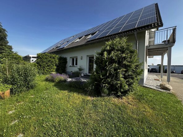 Residential house with a large array of solar panels on the sloped roof, front garden and driveway visible to the right.
