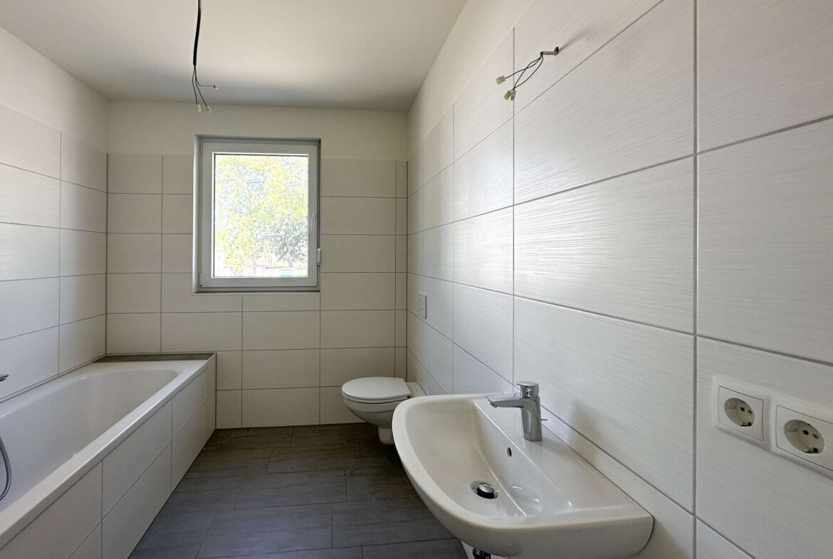 Modern bathroom with white tiled walls, bathtub on left, wall-mounted sink, and toilet under a small window; ceiling wires exposed, unfinished wiring visible.