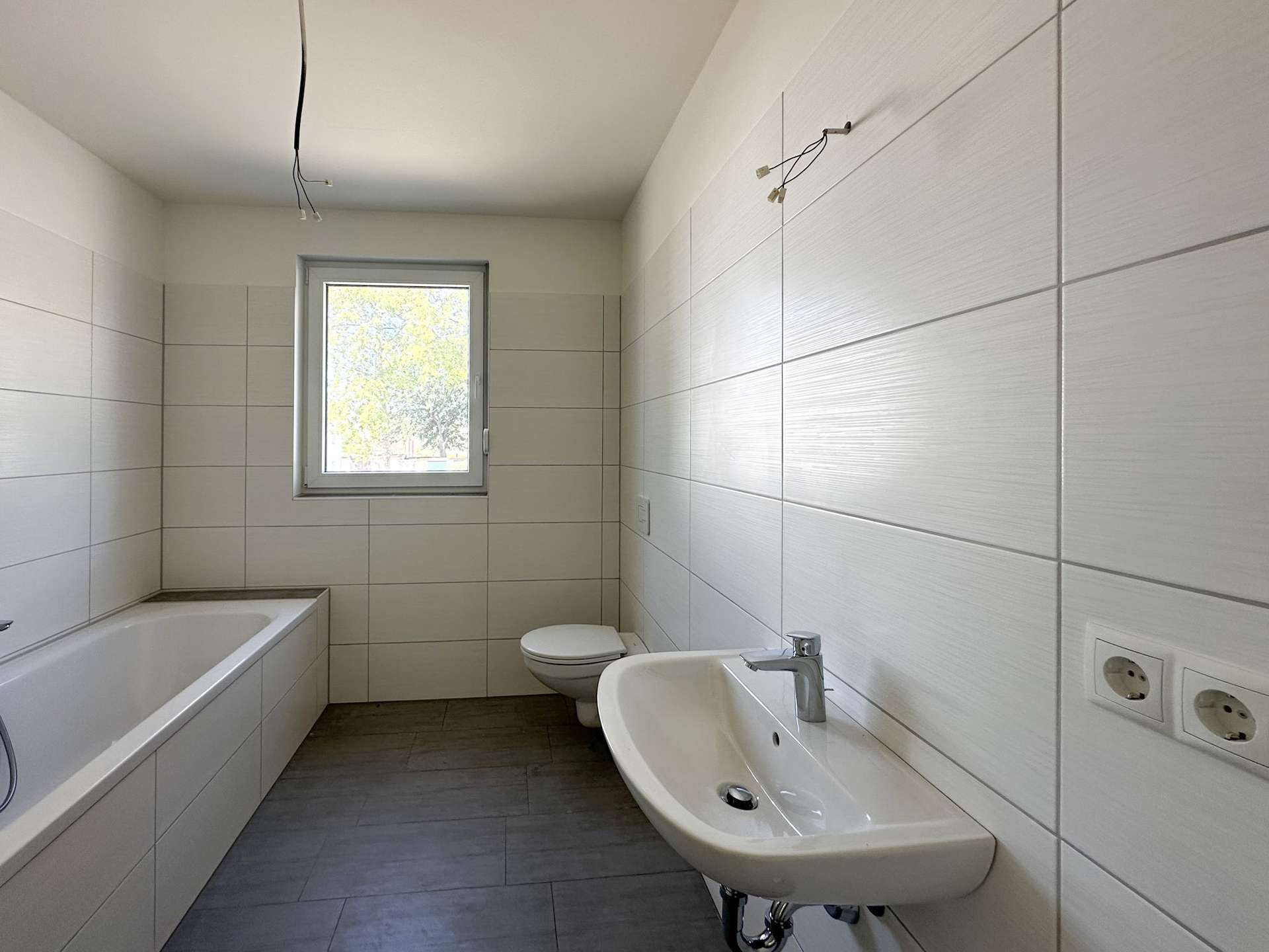 Modern bathroom with white tiled walls, bathtub on left, wall-mounted sink, and toilet under a small window; ceiling wires exposed, unfinished wiring visible.