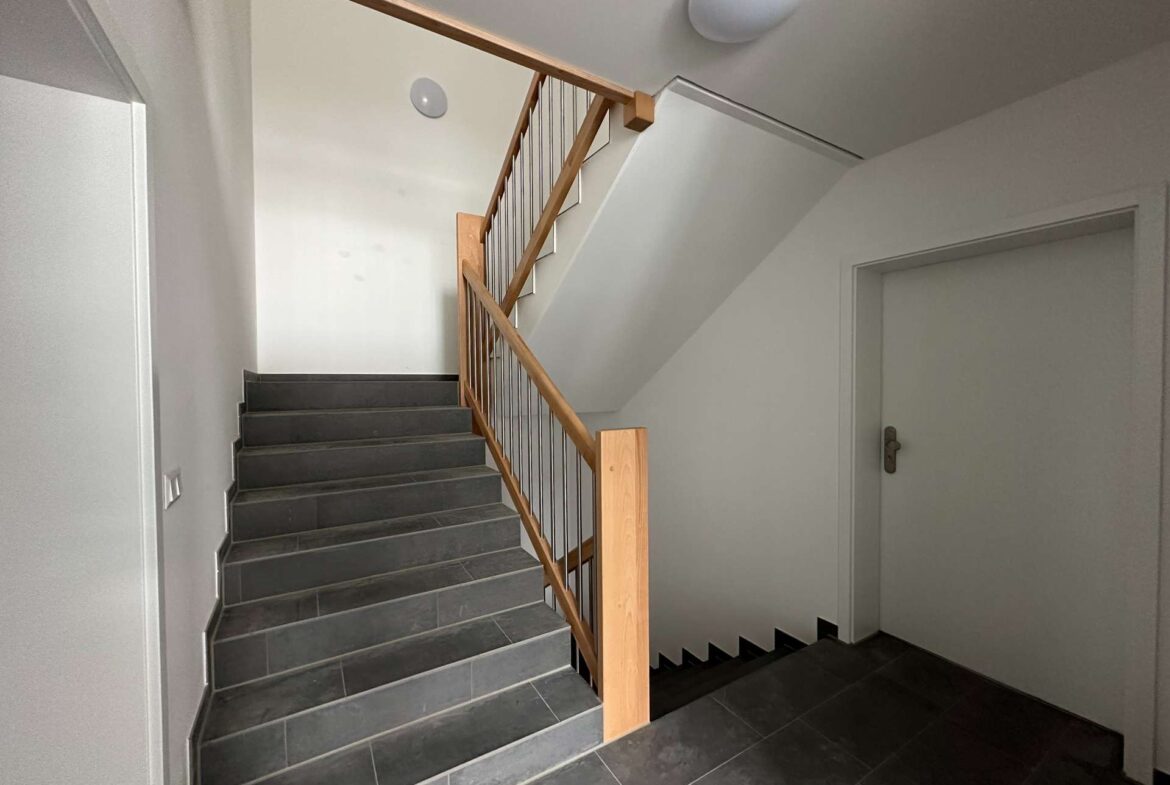 Interior stairwell with dark grey tile steps, wooden handrail and metal balusters, white walls, and a door to the right.