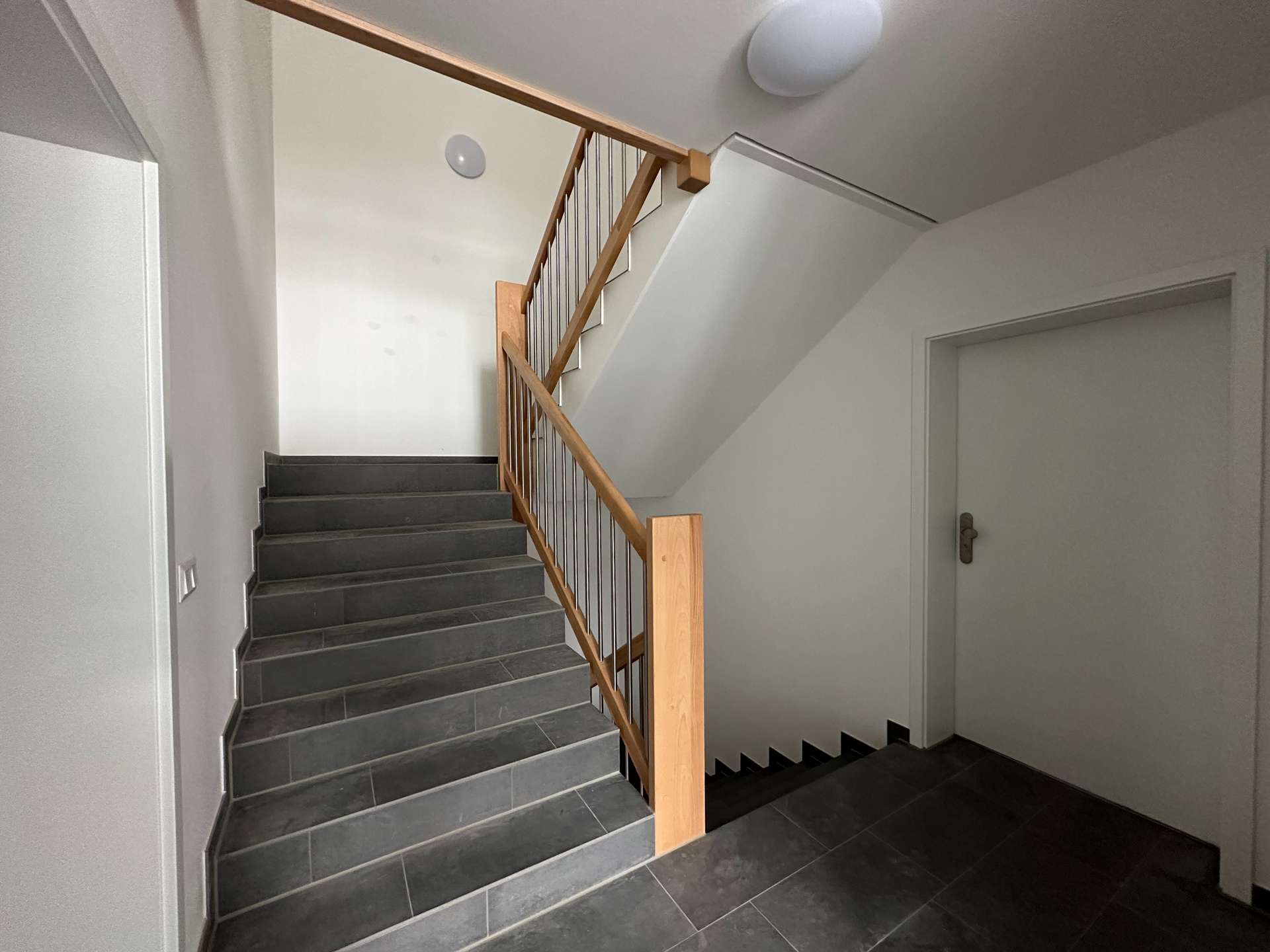 Interior stairwell with dark grey tile steps, wooden handrail and metal balusters, white walls, and a door to the right.