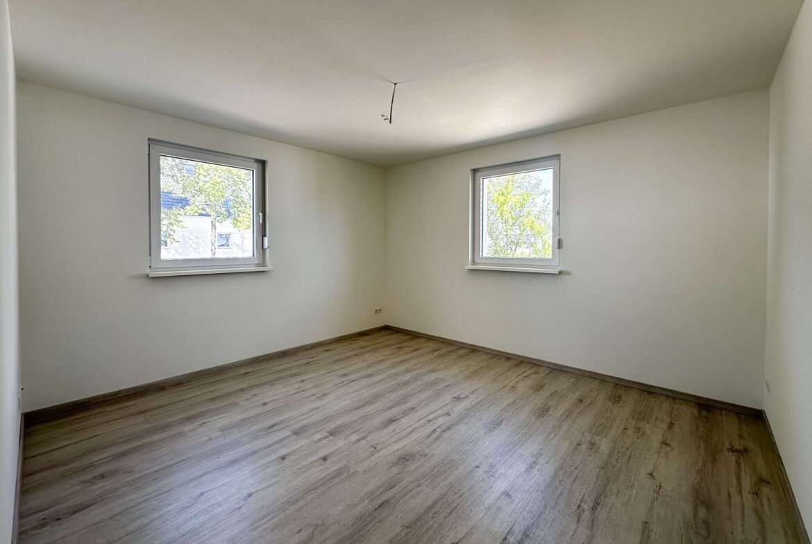 Empty rectangular room with two windows on opposite walls, light wood laminate flooring, white walls, and a hanging ceiling wire.
