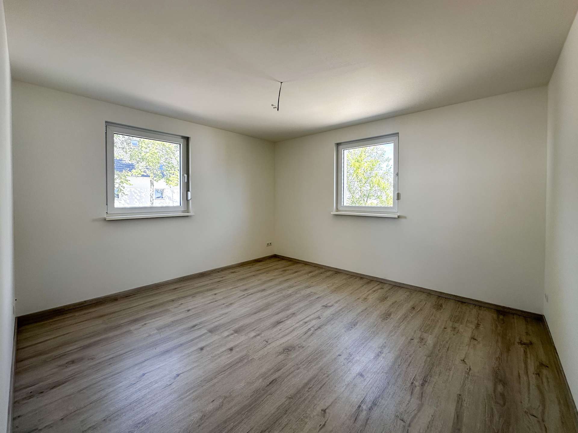 Empty rectangular room with two windows on opposite walls, light wood laminate flooring, white walls, and a hanging ceiling wire.