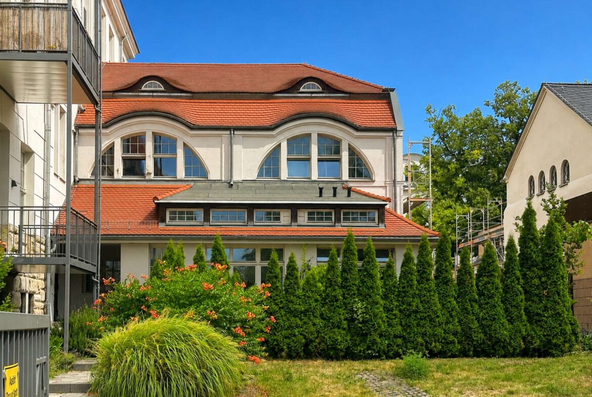 Cream-colored building with a red tiled roof and large arched windows, under a bright blue sky, with balconies on the left and greenery in front.