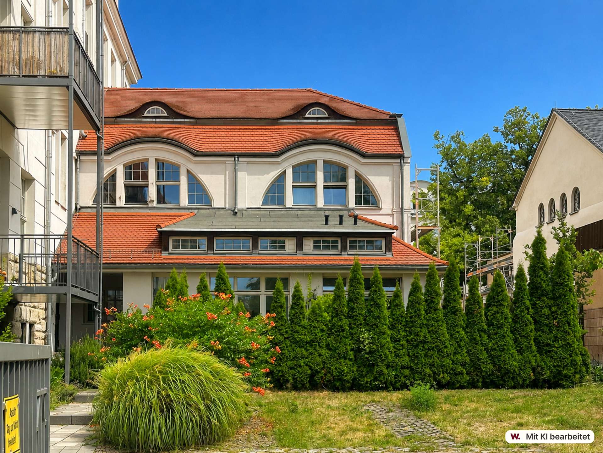 Cream-colored building with a red tiled roof and large arched windows, under a bright blue sky, with balconies on the left and greenery in front.