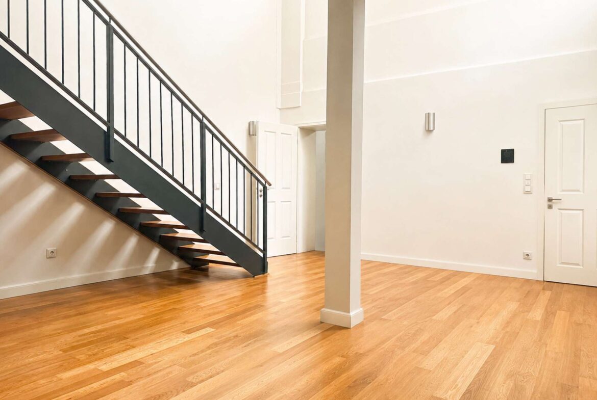 Open indoor staircase with dark metal railing and wooden steps along a pale wall, in a bright lobby with hardwood floors and a white door in the background.