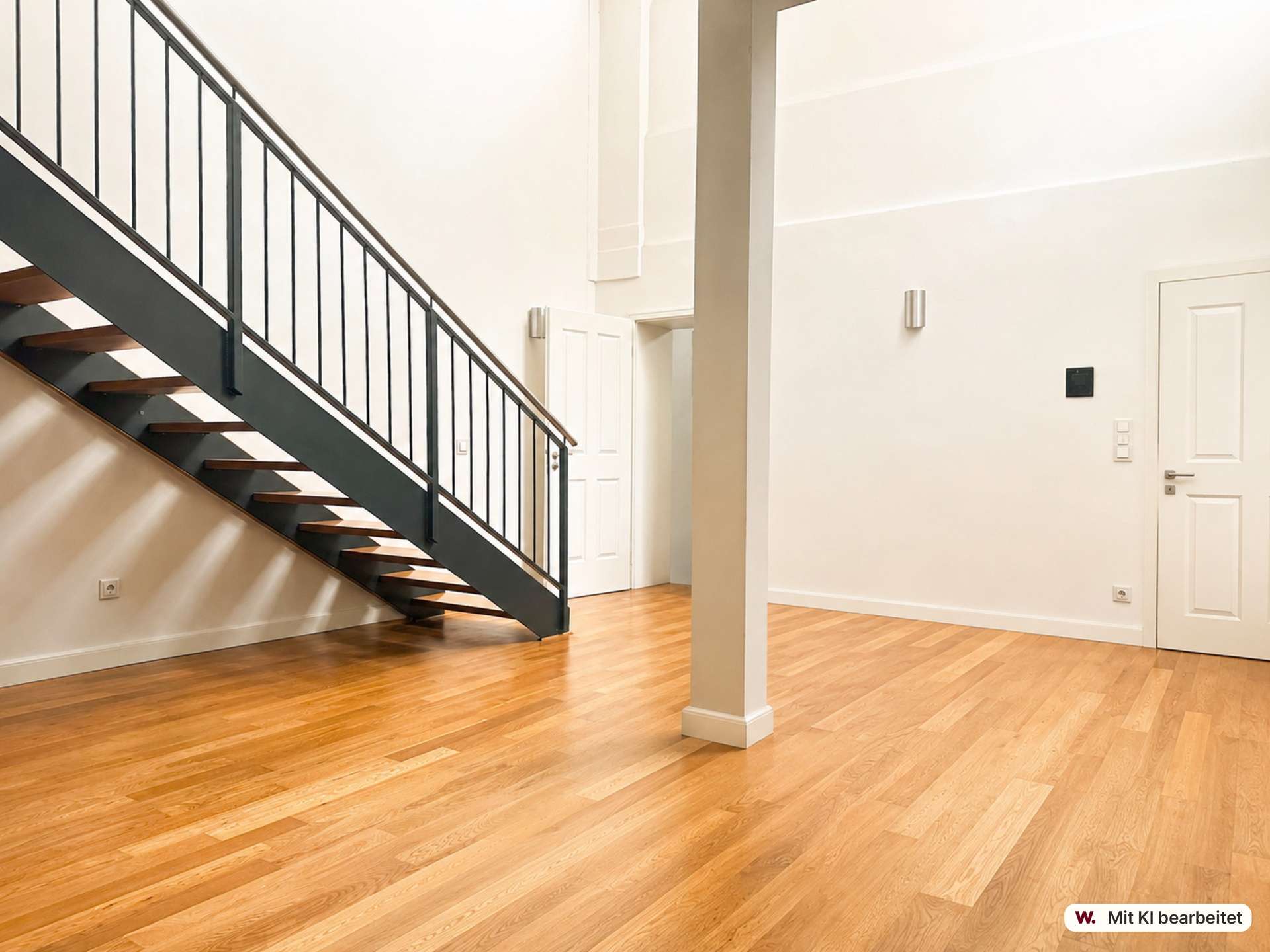 Open indoor staircase with dark metal railing and wooden steps along a pale wall, in a bright lobby with hardwood floors and a white door in the background.