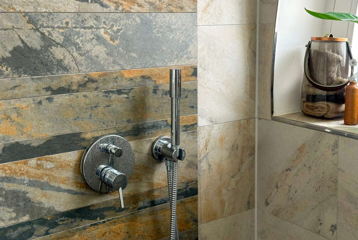 Modern shower wall with multitone stone tiles, a chrome handheld shower, and a towel-filled glass jar on the shelf nearby.
