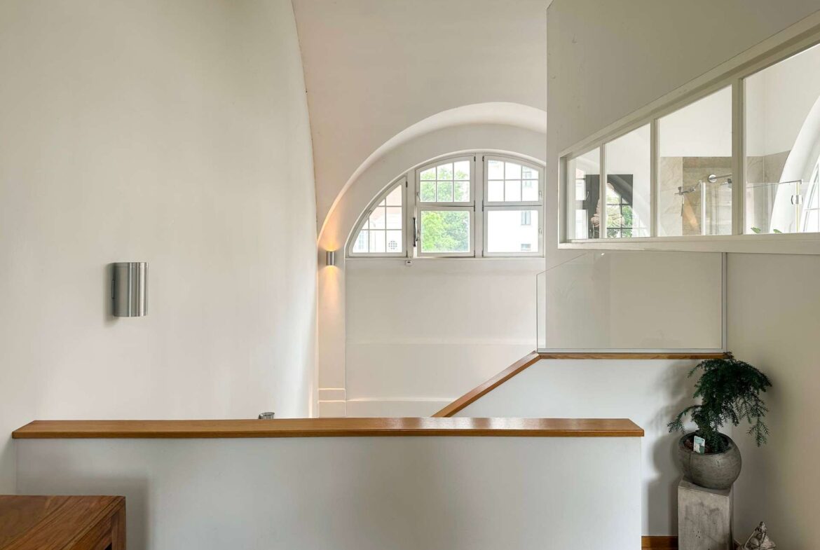 Interior hallway with a curved arch ceiling, large arched window, glass divider, wooden handrail, and a potted plant in the corner.