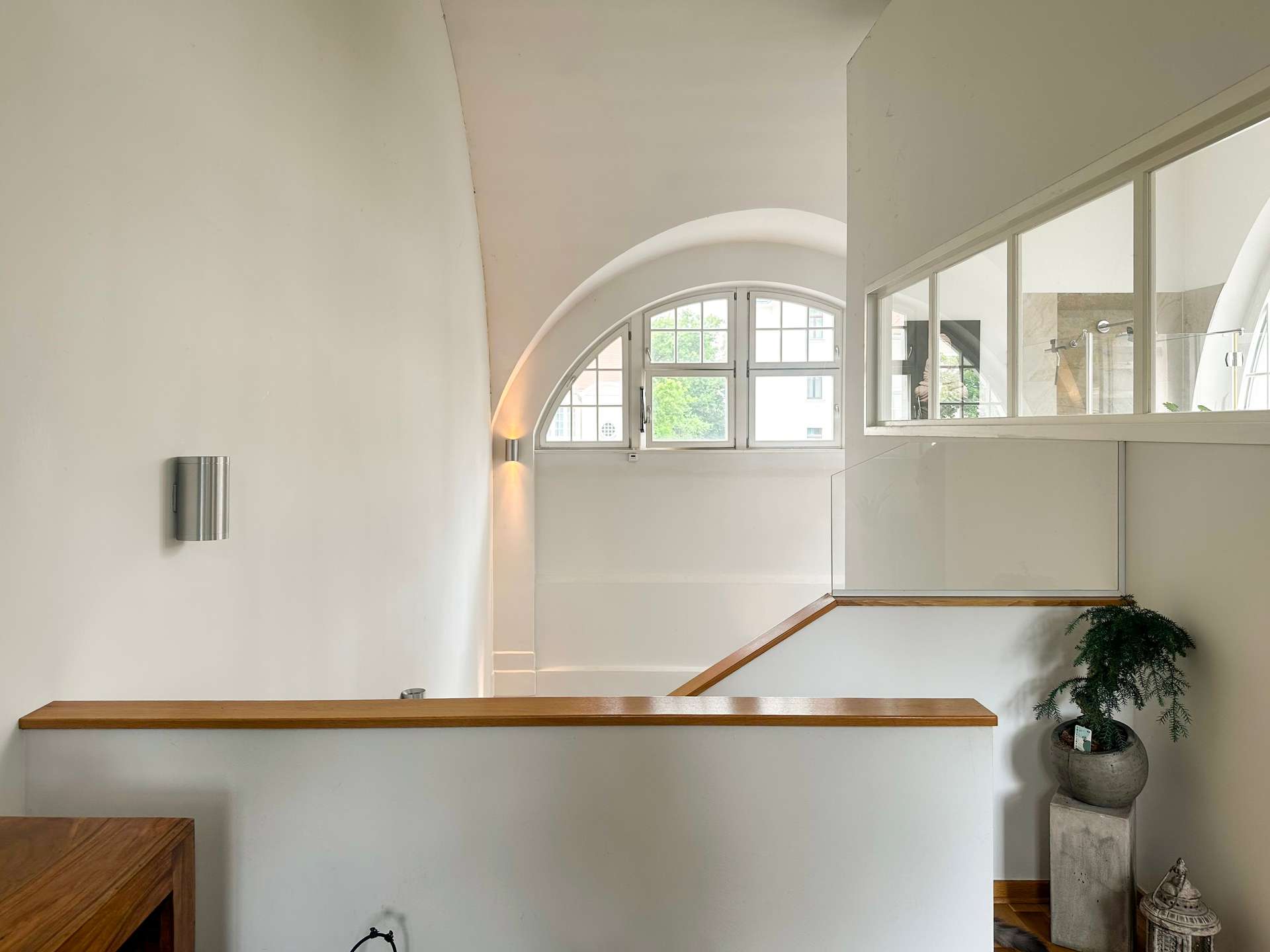 Interior hallway with a curved arch ceiling, large arched window, glass divider, wooden handrail, and a potted plant in the corner.