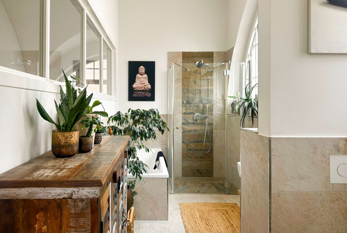 Bright, modern bathroom with a glass shower enclosure, beige tiled walls, and multiple potted plants on a rustic wooden cabinet beneath a Buddha wall art piece.