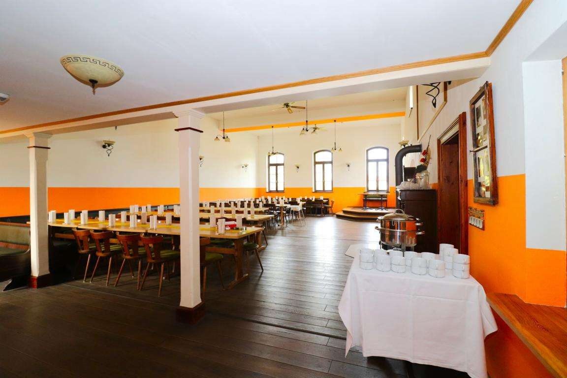 Communal dining hall with long wooden tables set with white cups and napkins, orange wainscoting, and a beverage station nearby.
