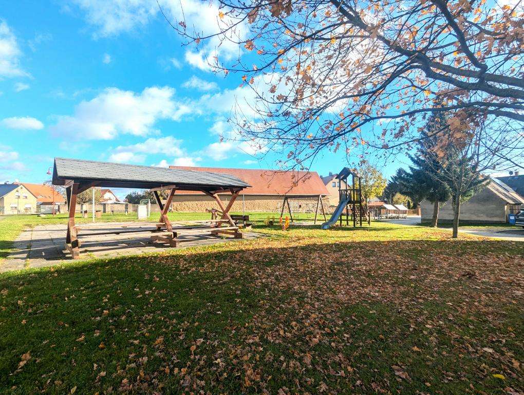 Park scene with a wooden picnic shelter and playground equipment on a sunny autumn day, leaves scattered on the grass.