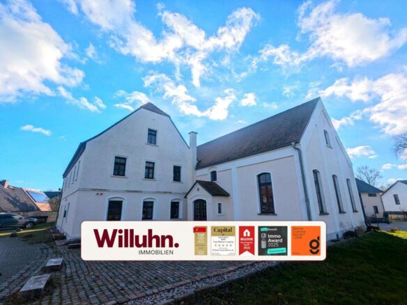 White multi‑wing building with a large Willuhn Immobilien banner and award badges in front under a blue sky.