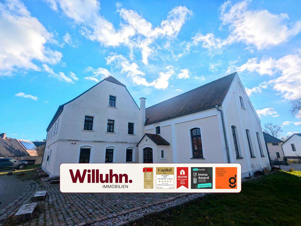 White multi‑wing building with a large Willuhn Immobilien banner and award badges in front under a blue sky.